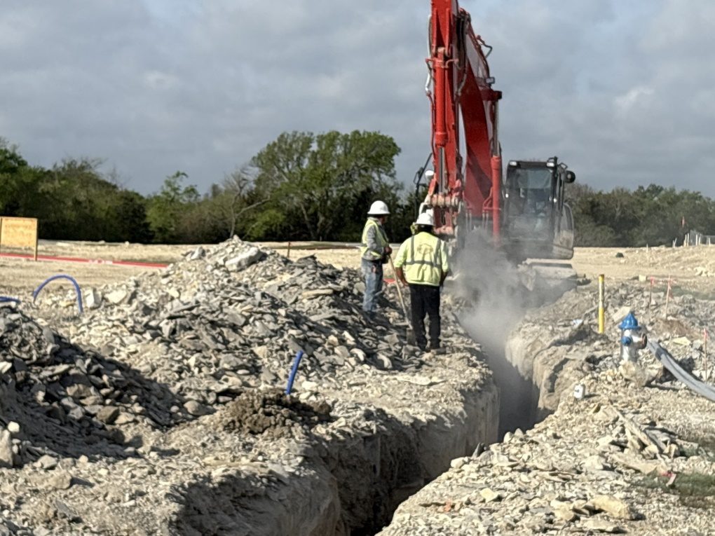 Crew watching a red excavator cut a rocky trench with dust in the air