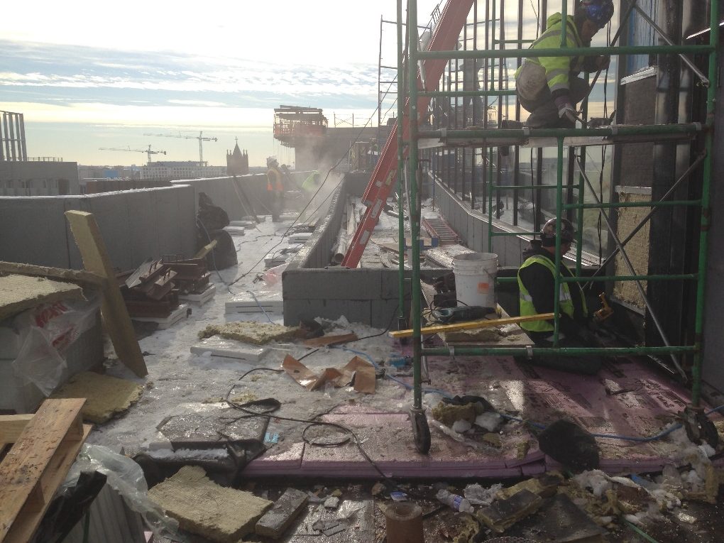 Workers on scaffolding at the edge of a messy high-rise deck with city and cranes beyond